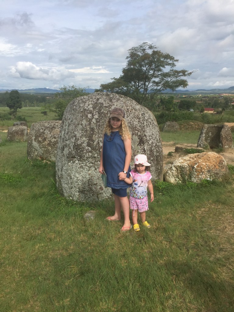 Two children standing next to a large megalith in a grassy area with trees and mountains in the background.