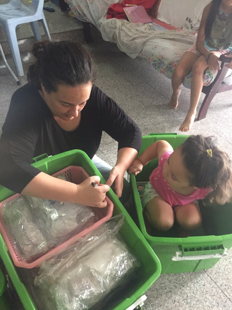 A woman in a black shirt organizes items in green storage bins while a young girl in a pink shirt sits in one of the bins, watching intently.